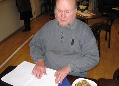 Bengt Eriksson sitter vid ett cafébord och läser punktskrift med  bägge händerna. Han har en grå tröja med krage och knappar i halsen. Bengt Eriksson har ljust skägg och tunt hår. Bredvid punktskriftstidningen ligger en leverpastejsmörgås med saltgurka. Foto: Åsa Nilsson
