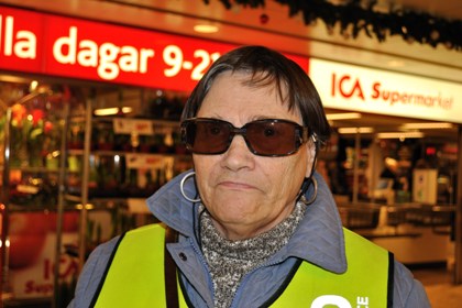 Lena Sahlsten har kort mörkt hår, mörka glasögon, stora runda örhängen. I bakgrunden en ICA-butik. Foto: Johan Rosengren.