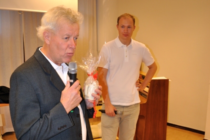 Björne Karlsson lämnar över en gåva till Olov Enström på distriktets medlemsforum. Olov Enström har en vit pikétröja och beiga jeans. Han har kort hår och ett högt hårfäste.