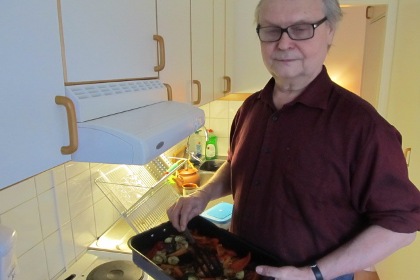 Guy Perdhe står i sitt kök. Han håller en form med grönsaksblandning i händerna. Han har grått bakåtkammat hår, är iklädd en mörkröd skjorta och bär glasögon.
