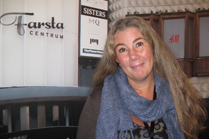 Tina Rodhe står framför en fototapet föreställande Farsta centrum. Tina Rodhe har långt brunt hår med grå inslag, och stora bruna ögon.