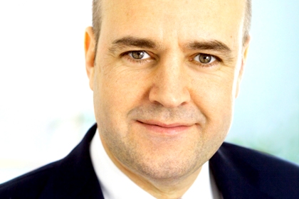 Fredrik Reinfeldt har kort mörkt hår på sidorna om huvudet, tydliga mörka ögonbryn, brungröna ögon, vit skjorta och mörkblå kavaj.
