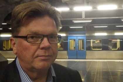 Lars Ericson har uppåtkammat hår, glasögon, bär kavaj och en blårandig skjorta. I bakgrunden ser man en tunnelbanevagn.