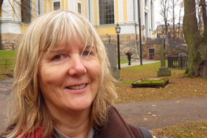 Elisabeth Brenning har axellångt ljust hår. Hon bär en brun kappa och en röd scarf.